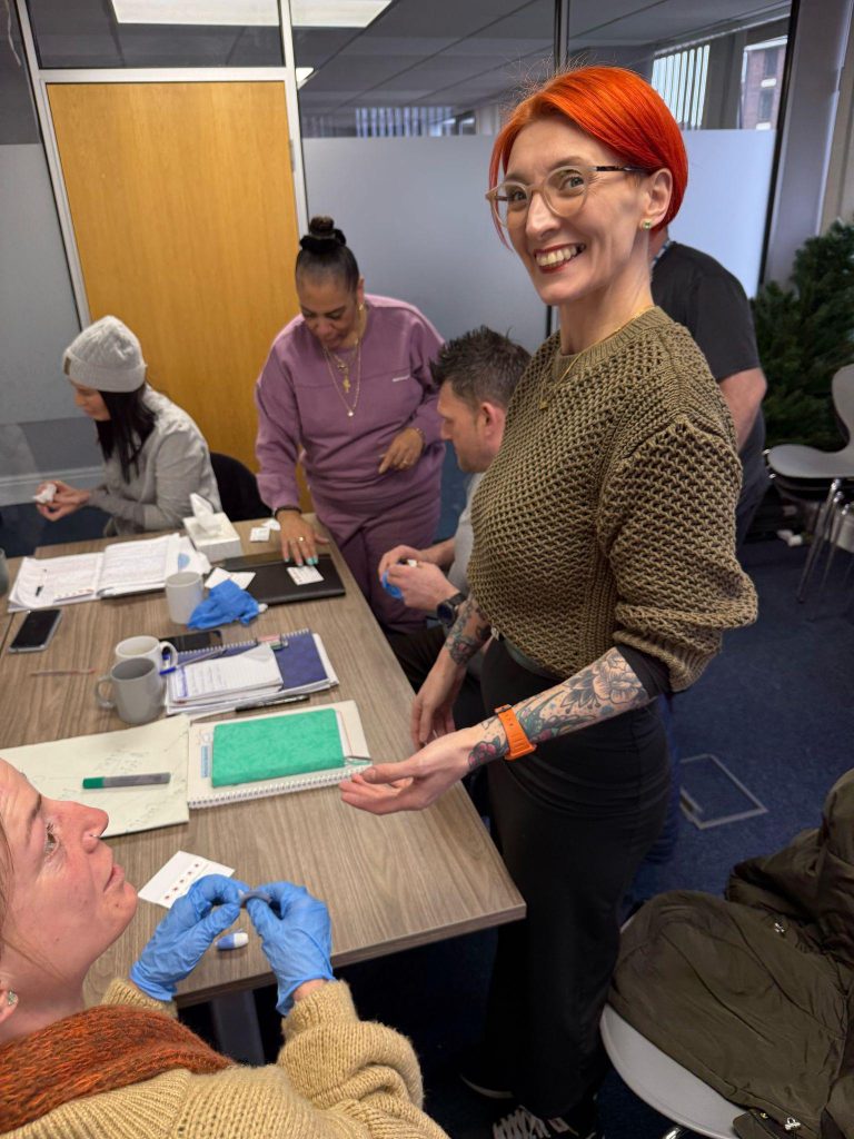 A group of five ambassadors stand around a table, you can see medical equipment on the table. Three of them are looking down at the table, working out how to test. One ambassador stands close to the camera with a big smile on her face.