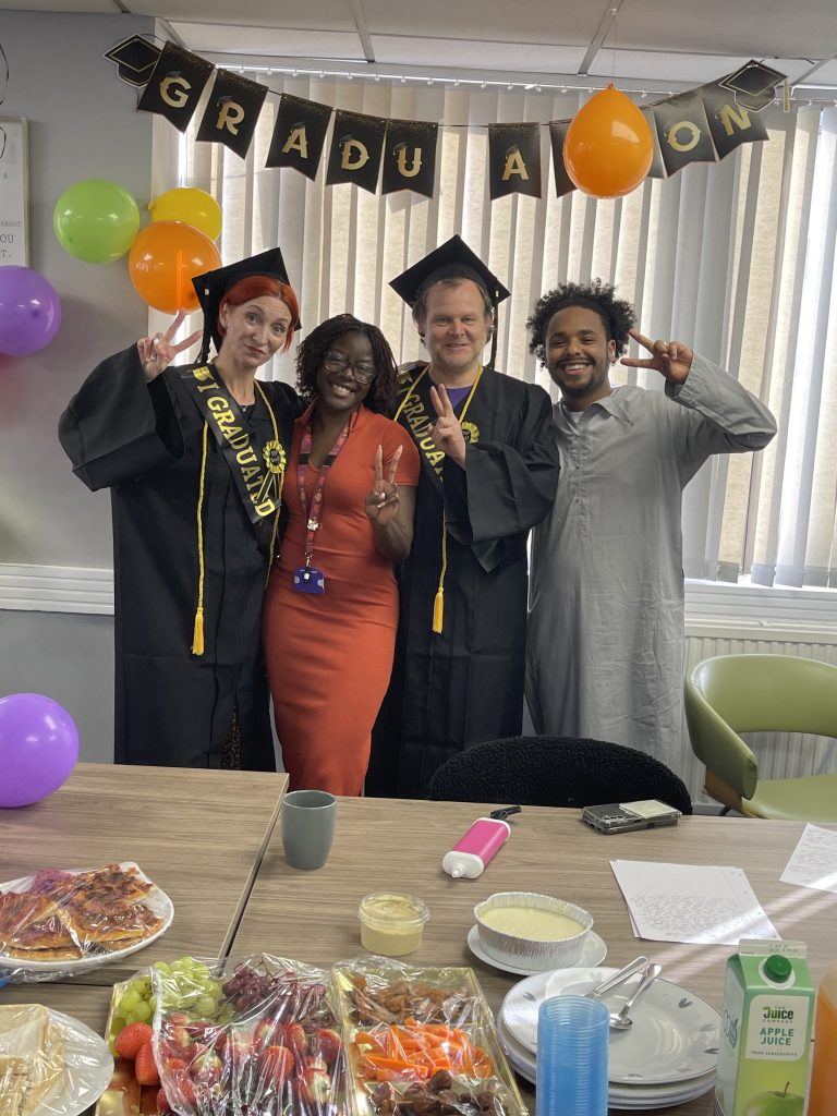 Two structured recovery graduates stand with the course faciltators, They are posing for the camera holding up peace signs. In front of them you can see party good laid out in the table. In the background there are graduate banner and balloons.