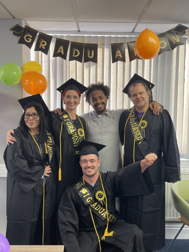 A group of five people stand together for a group photo. Four of them wear graduation gowns with black flat hats and golds sahses, while the man in the middle wear a plain clothes shirt. They all look very proud. In the background you can see graduation bunting and four baloon hanging from the ceiling.