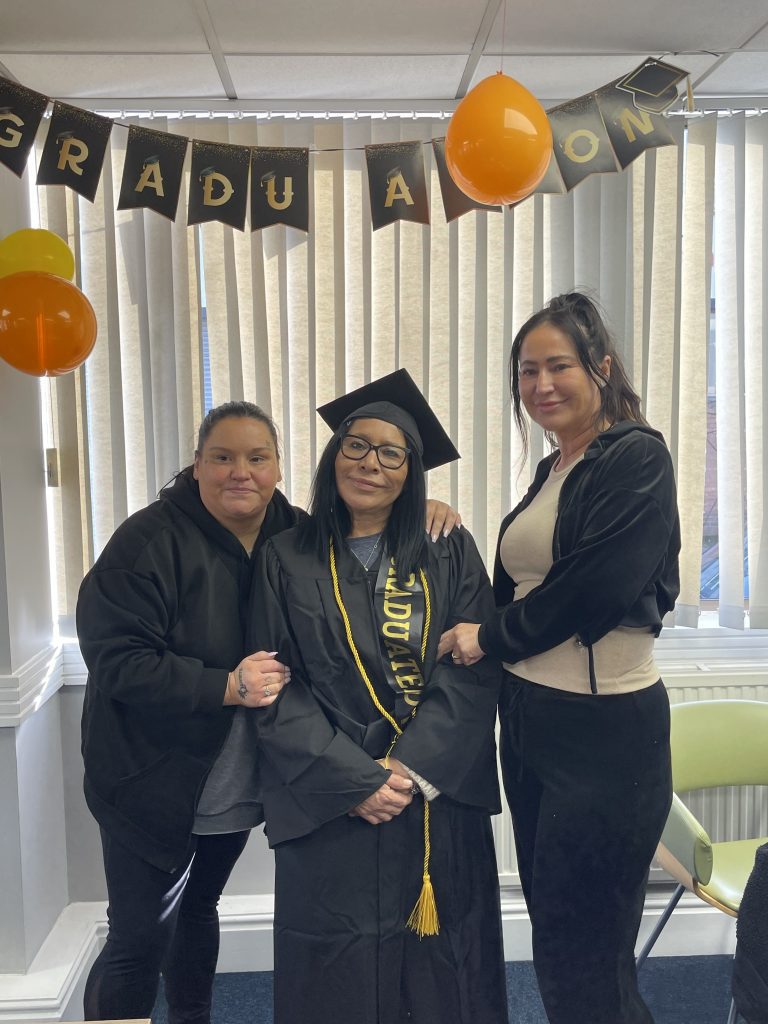 A photo of three women holding eachother arms. One woman stands in the middle in a graduation outfit wearing the flat cap, a gown and a gold sash. All three women are smiling into the camera. In the background you can see bunting that says 