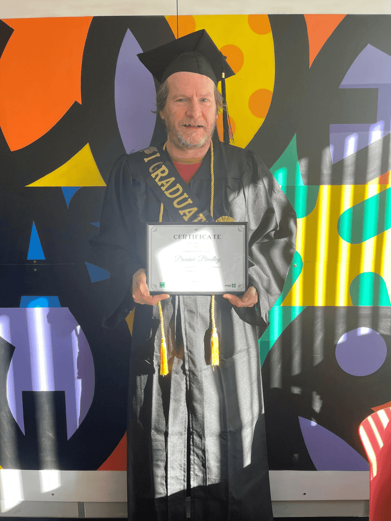 One of our SRP graduates stands wearing his graduation outfit, he is wearing a graduation gown and cap and sash that says graduate. He's holding a certificate. He stands against a colourful indoor mural.