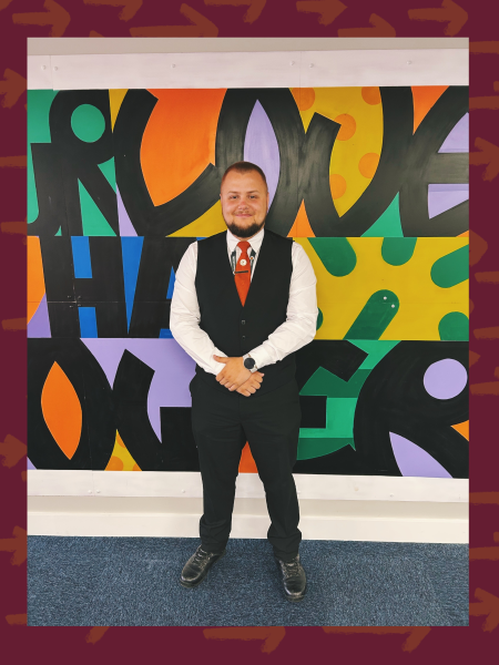 Portrait photo of a man wearing a security uniform, with a tie, white shirt and smart outfit. He stands in the centre of the photo smiling at camera, in background is a colourful mural with